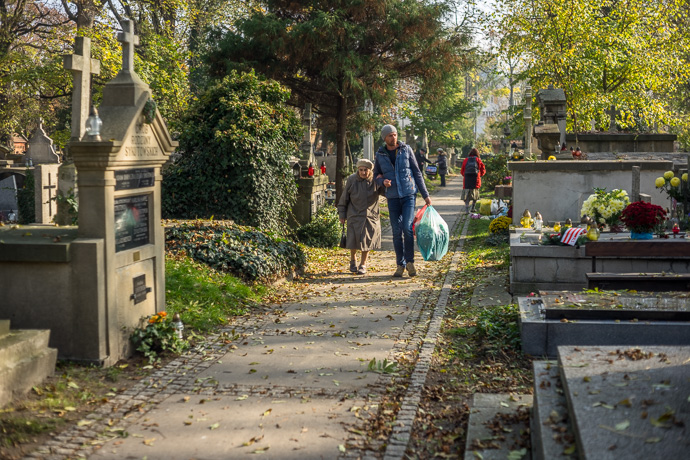 All Saints' Day at Rakowicki Cemetery in Kraków, Poland 2019. A man supports an old woman and carries plastic bags full of flowers as they walk through the cemetery, All Saints' Day in  Image: Michel Meijer